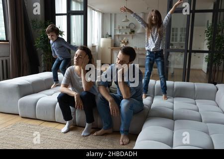 Laute Tochter und Sohn springen, müde Eltern sitzen auf der Couch Stockfoto