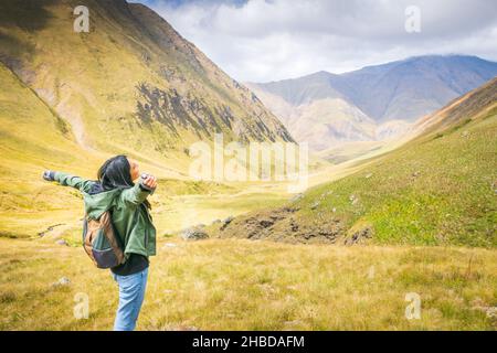 Die Backpacker-Frau steht auf dem Felsen und genießt einen atemberaubenden Blick auf die Berge im Juta-Tal. Erkundung des Nationalparks Kazbegi Stockfoto