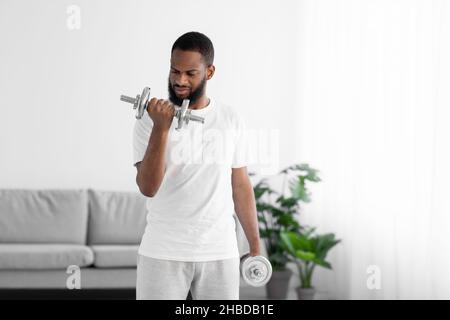 Seriöse junge schwarze Sportler Mann in weißen Kleidern tun Übung mit Hanteln im minimalistischen Wohnzimmer Stockfoto