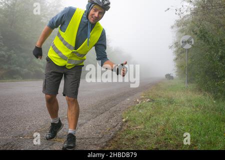 Ein glücklicher Radfahrer mit einer reflektierenden Weste zeigt bei nebligen Bedingungen die Daumen nach oben auf der Straße mit einem Touring-Fahrrad in der Nähe. Konzept für sicheres Radfahren unter b Stockfoto