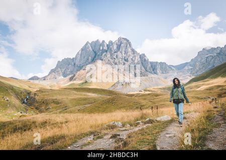 Glückliche weibliche Kaukasierin geht den Weg im Juta-Tal mit landschaftlich reizvoller Berglandschaft hinunter. Reiseziel Kazbegi Stockfoto