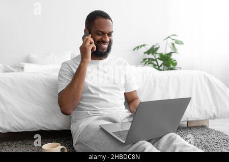 Lächelnder, schwarzer bärtiger Mann in weißer Kleidung sitzt auf dem Boden, arbeitet am Laptop, spricht auf dem Smartphone Stockfoto