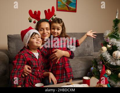 Glückliche Mutter mit Geweih-Reifen umarmt ihre entzückenden Kinder- Sohn und Tochter in rot und grün karierten Kleidern und Weihnachtsmann Hut auf dem Kopf ihres Sohnes, Celebra Stockfoto