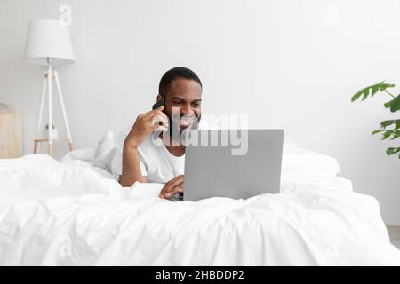 Lächelnder junger, schwarz bärtiger Mann liegt auf dem Bett in einem weißen, minimalistischen Schlafzimmer und spricht per Telefon Stockfoto