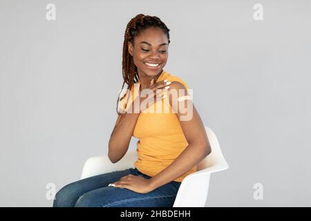 Tausendjährige schwarze Frau, die gegen Kovid geimpft wird, zeigt einen Arm mit Verband auf grauem Studiohintergrund Stockfoto