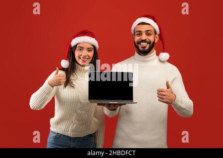 Mockup Bild von glücklichen arabischen Paar in Santa Hüte halten Laptop mit leerem Bildschirm und zeigt Daumen nach oben Stockfoto