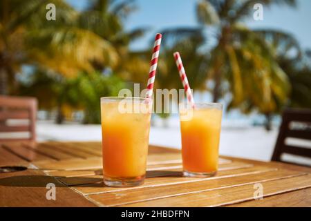 Zwei erfrischende kalte Getränke am weißen Sandstrand. Gläser Cocktails mit Trinkhalmen aus Papier auf einem Holztisch gegen Palmen. Stockfoto