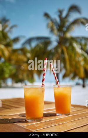 Zwei erfrischende kalte Getränke am weißen Sandstrand. Gläser Cocktails mit Trinkhalmen aus Papier auf einem Holztisch gegen Palmen. Stockfoto