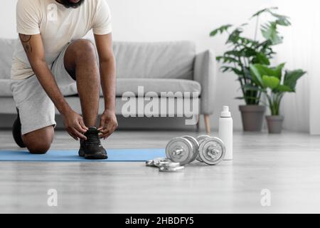 Der kurzgeschnittene junge schwarze Athlet Mann in weißer Sportswear bindet Schnürsenkel an Schuhe und bereitet sich auf die Übungen vor Stockfoto