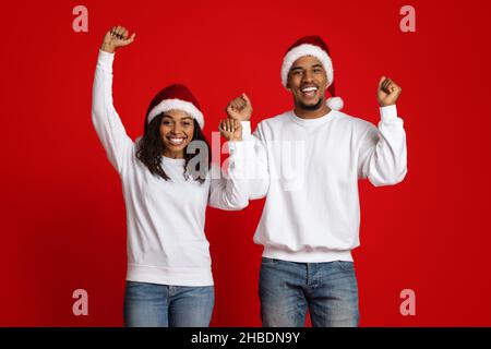 Fröhliches schwarzes Paar in Weihnachtsmützen, gestikuliert auf Rot Stockfoto