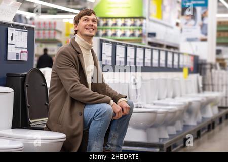 Mann mit brünettem welligen Haar in braunem Mantel und warmem Wollpullover wählt Toilette für Wohnungsrenovierung sitzend und lächelnd im Supermarkt Stockfoto