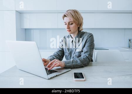 Eine Geschäftsfrau in einem grauen Hemd mit kurzem Haarschnitt arbeitet an einem Laptop, während sie an einem Holztisch in einem hellen Büro sitzt, steht ein Arbeitstelefon auf dem Tisch Stockfoto