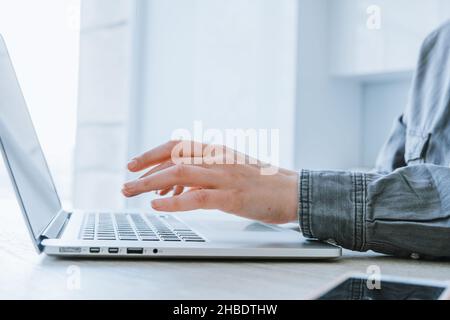Hände einer Geschäftsfrau in einem grauen Hemd, die hinter einem Laptop in einem hellen Büro auf dem Hintergrund arbeitet, aus der Nähe Stockfoto