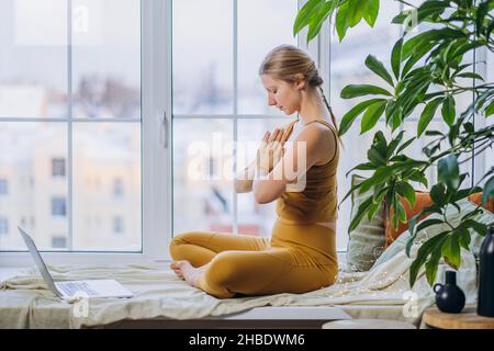 Die junge blonde langhaarige Frau in gelber Farbe meditiert in Lotuspose und sitzt auf einer Decke, die das Fensterbrett bedeckt, und schaut auf den Laptop-Bildschirm Stockfoto