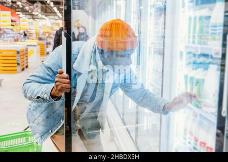 Afrikanischer Hipster-Mann öffnet die Glastür eines Kühlschranks in einem Supermarkt und wählt frische abgefüllte Milch oder Kefir und Joghurt Stockfoto