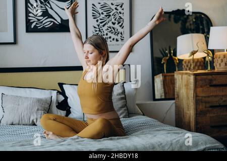 Die junge blonde langhaarige Frau im Sporttop und in Leggings streckt sich auf dem Bett sitzend, das mit einer Decke und weichen Kissen bedeckt ist, gegen den Spiegel Stockfoto