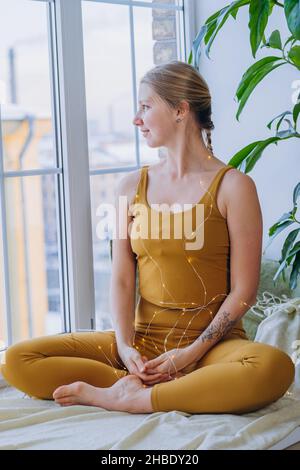 Frau mit blondem Geflecht, das ein gelbes Oberteil trägt und Leggings, die mit leuchtendem Girlande umhüllt sind, genießt den Blick auf die Stadt vor dem Fenster, das auf dem Fensterbrett sitzt Stockfoto