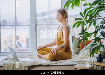 Die blonde junge Frau in gelbem Oberteil und Leggings meditiert in Yoga-Pose und sitzt auf einem mit Decke und Kissen bedeckten Fensterbrett mit einem Laptop Stockfoto