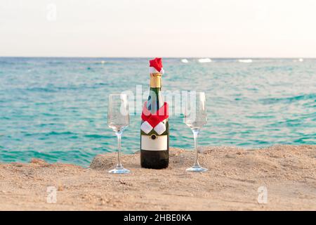 Eine fröhliche Flasche Champagner in einem Weihnachtsmannhut und zwei Gläser am Strand vor dem Hintergrund des Meeres. Weihnachtskonzept. Stockfoto
