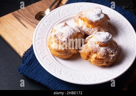 Lebensmittelkonzept hausgemachte frisch gebackene Choux a la Creme, Cream Puff, Choux Cream in weißer Keramikplatte auf schwarzem Hintergrund mit Kopierfläche Stockfoto