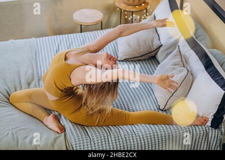Die junge blonde Frau trägt ein Sporttop und Leggings, die Yoga-Posen machen, und dehnt sich auf einer weichen Decke, die das Bett mit Girlande-Beleuchtung bedeckt Stockfoto