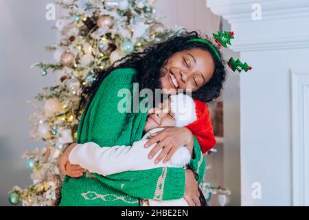 afroamerikanische Mama in grünem Pullover und Stirnband mit weihnachtsbäumen umarmt ihre kleine Tochter in einem weihnachtshut vor dem Hintergrund eines weihnachtsbaums. Stockfoto