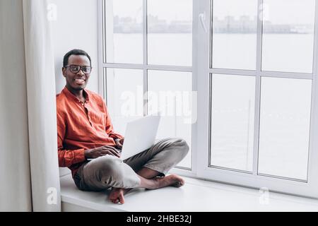 Ein junger afrikanischer Geschäftsmann mit Brille sitzt auf einem Laptop auf dem Fensterbrett eines Hauses und schaut lächelnd auf die Kamera Stockfoto