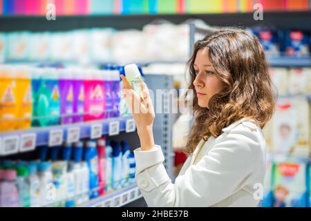 Brünette Frau mit langen lockigen Haaren trägt weißen Mantel liest Anweisung auf Deodorant-Paket stehen im Haushalt Waren speichern Stockfoto