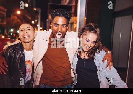 Drei junge Freunde, die abends in der Stadt Spaß haben. Eine Gruppe von jungen Menschen, die beim gemeinsamen Gehen fröhlich lachen. Multikulturelle Freunde gehen Stockfoto