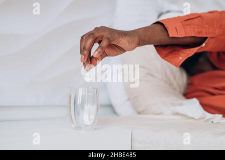Kranker Afrikaner im Schlafanzug wirft zwei Fiebertabletten in ein Glas Wasser, Hand in Nahaufnahme. Behandlung von Influenza oder Coronavirus, Krankenhausaufenthalt und Bettruhe. Stockfoto
