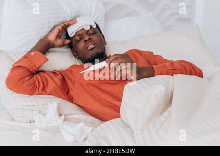 Der kranke Afrikaner im orangefarbenen Pyjama misst die Temperatur mit einem elektronischen Thermometer, während er mit einer Kühlkompresse auf dem Kopf im Bett liegt. Schwarzer Mann hat Fieber und leidet unter dem Coronavirus. Stockfoto