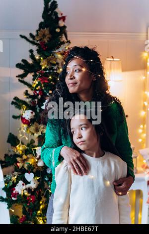 Brünett Mädchen und lange lockige afroamerikanische Mutter Pose mit leuchtenden Girlande auf dem Kopf und lächeln glücklich gegen Weihnachtsbaum Stockfoto