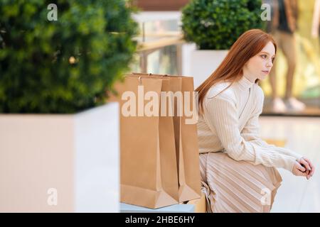 Müde hübsche junge Frau, die mit Papiertaschen auf der Bank im Einkaufszentrum mit moderner Innenausstattung und verschwommenem Hintergrund sitzt. Stockfoto