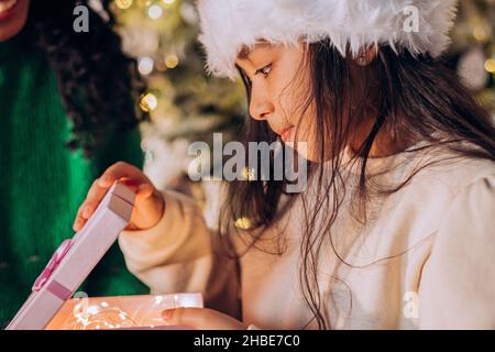 Brünette langhaarige Tochter in Santa Hut öffnet festliche Box mit Geschenken sitzen in der Nähe afroamerikanische Mutter gegen Weihnachtsbaum Stockfoto