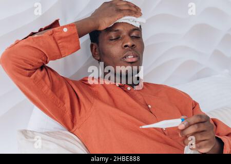 Der kranke Afrikaner im orangefarbenen Pyjama misst die Temperatur mit einem elektronischen Thermometer, während er mit einer Kühlkompresse auf dem Kopf im Bett liegt. Schwarzer Mann hat Fieber und leidet unter dem Coronavirus. Stockfoto