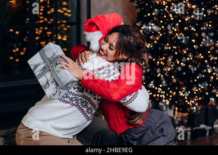 Afroamerikanisches Paar umarmungmit Neujahr Geschenk auf dem Hintergrund von weihnachtsbaum, Atmosphäre der Feier und Familie Glück, junge schöne Paar in gemusterten Pullover Stockfoto