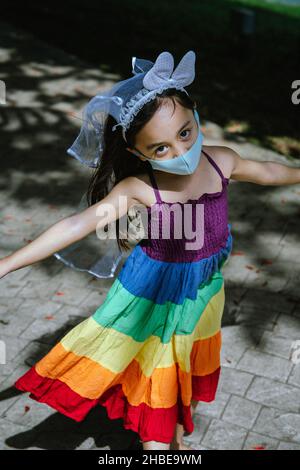 Kleines Mädchen trägt Gesichtsmaske mit Regenbogenkleid genießen im Freien im Park. Neuer Normalwert. Stockfoto