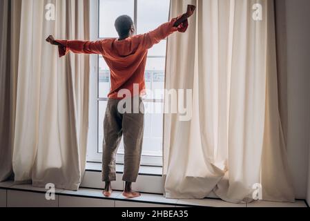 Afrikanischer Mann in orangefarbenem Pyjama und Hose streckt sich nach dem Aufwachen am Fenster stehend mit Vorhängen und blickt in die Ferne, macht Morgenübungen, den Beginn eines neuen Tages Stockfoto