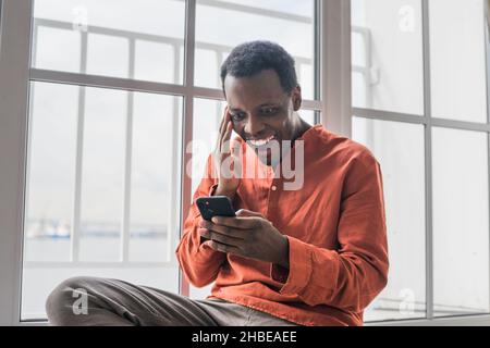 Glücklich überrascht afroamerikanischen Mann in stilvollen orange Hemd sieht auf Smartphone-Bildschirm sitzen Fensterbank im Zimmer zu Hause Stockfoto