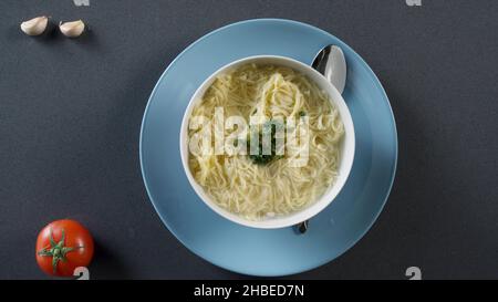 Hühnersuppe mit Nudeln auf einem Teller. Hühnernudelsuppe in cremefarbener Keramikschale mit Griffen. Teller mit Crackern und Suppenschüssel im Hintergrund Stockfoto