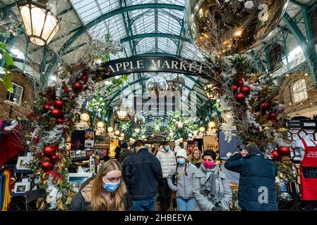 London, Großbritannien. 19. Dezember 2021. Menschen, die am letzten Einkaufswochenende vor Weihnachten Gesichtsmasken unter den Dekorationen auf dem Apple Market von Covent Garden tragen. Die Fälle der Omicron-Variante sind so stark angestiegen, dass die britische Regierung mit der Umsetzung strengerer Plan-C-Beschränkungen konfrontiert ist, um ihre Ausbreitung einzudämmen, wie zum Beispiel einem Verbot der Vermischung in Innenräumen zwischen Haushalten und einem Verbot der Innenbewirtung. Kredit: Stephen Chung / Alamy Live Nachrichten Stockfoto