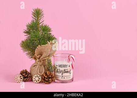 Weihnachtsbaum in Jutebeutel mit Tannenzapfen, Kerze mit dem Aufdruck „Frohe Weihnachten“ und Schneeflocke-Ornament auf rosa Hintergrund mit Kopierraum Stockfoto