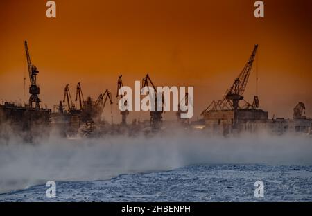 Der Bau von nuklearen Eisbrechern bei magischem Sonnenuntergang, Kräne der baltischen Werft in einem frostigen Wintertag, Dampf über dem Fluss Neva, glatt Stockfoto