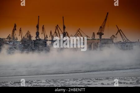 Der Bau von nuklearen Eisbrechern bei magischem Sonnenuntergang, Kräne der baltischen Werft in einem frostigen Wintertag, Dampf über dem Fluss Neva, glatt Stockfoto