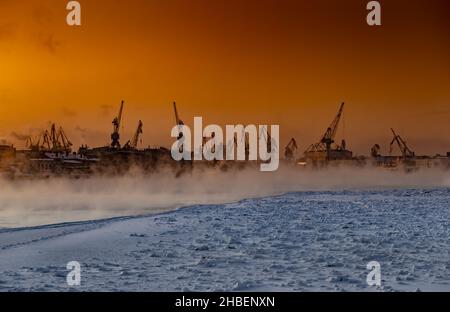 Der Bau von nuklearen Eisbrechern bei magischem Sonnenuntergang, Kräne der baltischen Werft in einem frostigen Wintertag, Dampf über dem Fluss Neva, glatt Stockfoto