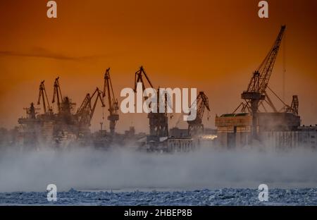 Der Bau von nuklearen Eisbrechern bei magischem Sonnenuntergang, Kräne der baltischen Werft in einem frostigen Wintertag, Dampf über dem Fluss Neva, glatt Stockfoto