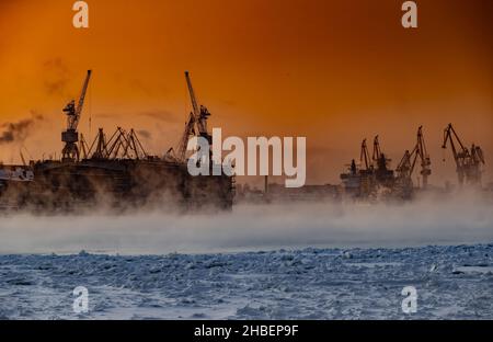 Der Bau von nuklearen Eisbrechern bei magischem Sonnenuntergang, Kräne der baltischen Werft in einem frostigen Wintertag, Dampf über dem Fluss Neva, glatt Stockfoto