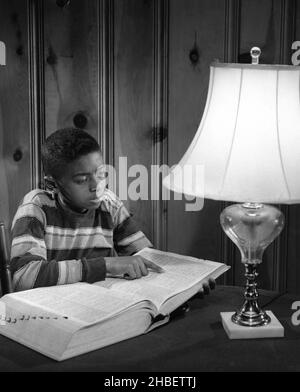 Afroamerikanischer Junge, der vor einer großen Lampe sitzt und ein großes Buch offen hat und auf Seite zeigt Stockfoto