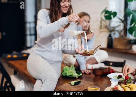 Die junge Frau sitzt auf dem Küchentisch und gießt Milch in Müsli Stockfoto