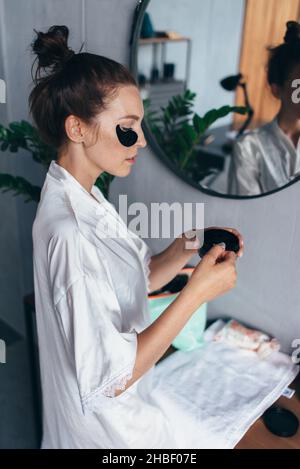 Die junge Frau nimmt Flecken aus einem Glas und trägt sie unter ihren Augen auf. Stockfoto